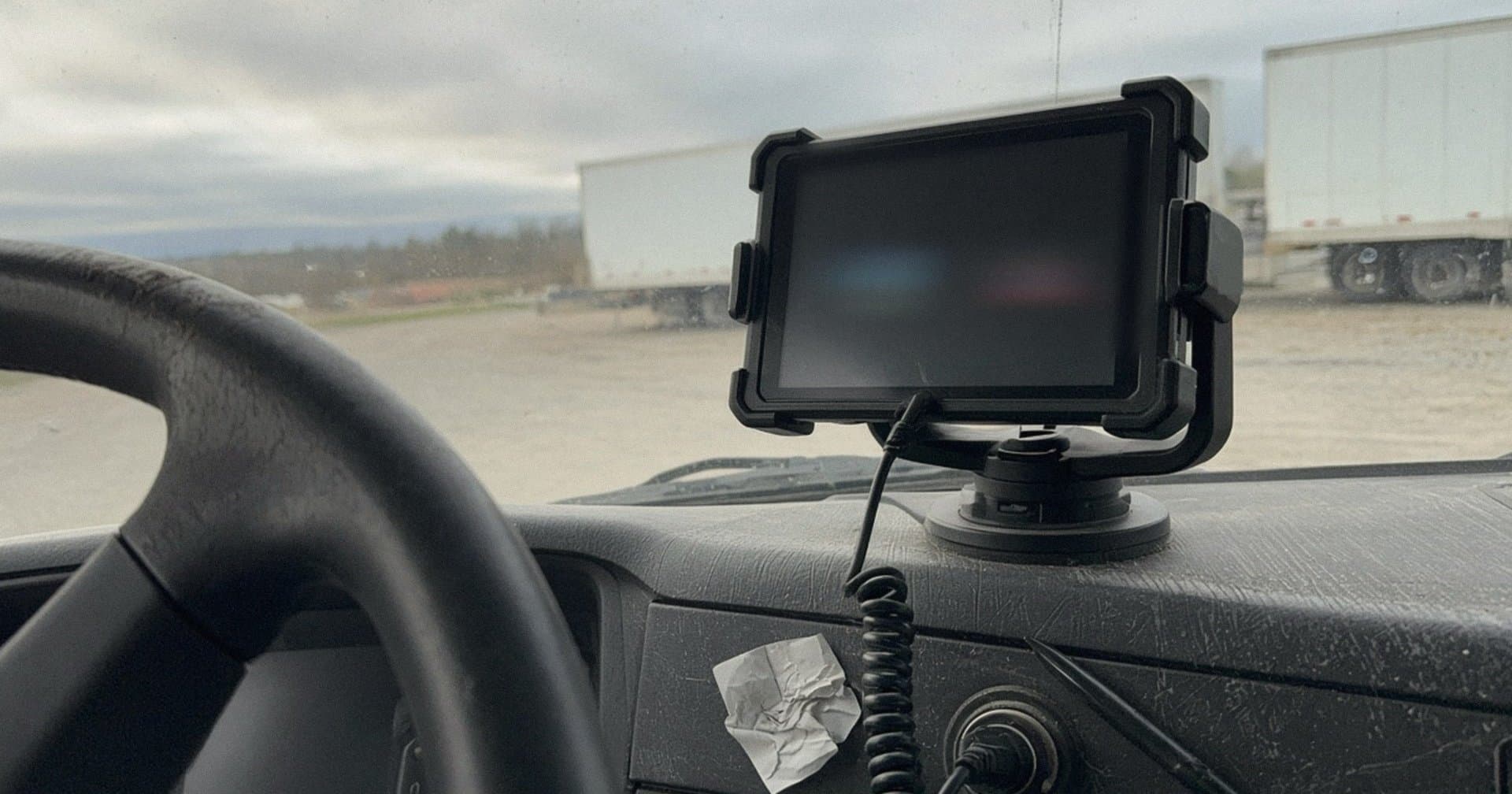 Dash-mounted electronic logging device in a rugged cradle with a coiled cable, inside a semi truck cab parked at a truck lot on an overcast morning